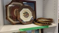 Shelf display of stacked frames showing various shapes: round gilded frames and octagonal wood and gold frames with interior needlepoint designs