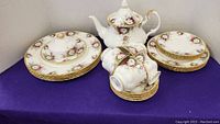 Overview on purple cloth showing teapot, covered butter dish, stack of dinner and luncheon plates, and cups with saucers