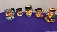 Front view of all five Toby jugs displayed in a row on a purple surface showing various characters and vibrant colors.