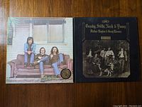 Two vinyl albums laying flat on a wooden surface showing front covers. Left album is Crosby, Stills & Nash with three band members seated on a couch. Right album is Deja Vu by Crosby, Stills, Nash & Young with a sepia toned group photo and gold text.