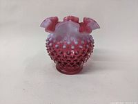 Front and angled view of cranberry colored glass vase with hobnail texture and ruffled rim on neutral background.