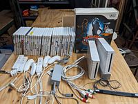 Two Nintendo Wii consoles, one standing vertically, one laid flat, with multiple game cases lined up behind them, various accessories including controllers, sensor bar, and power adapter.