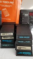 Eight Atari 2600 game cartridges stacked showing titles including Lock 'N' Chase, Tron Deadly Discs, Dark Cavern, Frogs and Flies, Adventures of Tron, Star Raiders, Astrobalt, Space Attack, and Super Challenge Baseball with a Zoom 505 Guitar controller and an orange boxed Video Touch Pad in the background.