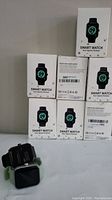 Eight boxes of smart watches labeled 'Smart Watch Your Sports Partner' stacked in two columns. Two watches with black and green silicone bands displayed in front showing square digital screens.