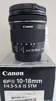 Canon EFS 10-18mm lens mounted vertically showing focus and zoom rings, manual/auto focus switch, marked focal lengths. Lens placed on original Canon box which shows the lens model details.