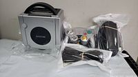 Nintendo GameCube console standing vertically with handle, silver color, in plastic wrap, alongside silver wired controller, power supply, and cables.