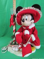 Front angled view of Minnie Mouse figure in red holiday clothing with candy cane lamp post base wrapped in garland and bow. Showing details of figure, base, and attached cord.