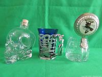 Three glass barware items set against a green cloth: a skull-shaped bottle, cobalt blue glass stein with silver overlay, clear glass stein with ceramic lid.