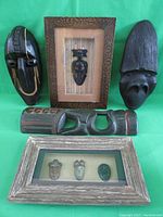 Overall view of the five carved wood items against green background, including masks and framed art