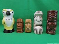 Five items arranged side by side on green background: yellow parrot-shaped shaker, two small tiki mugs (brown and tan), one tall cream tiki mug with vertical brown lines, and one brown tiki salt and pepper shaker with carved face design.