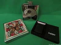 Photo showing the black matte Zippo lighter in its box, a shiny metal flask, and a vintage decorated metal case with butterfly design.