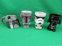 Four Star Wars character mugs from left to right: classic Boba Fett helmet, stylized Boba Fett mug, Stormtrooper helmet, and Darth Vader helmet mug, all ceramic with detailed paint and sculpt.