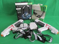 Two boxed Xbox 360 headphones and laser tag game set with two white laser tag guns, vests, and a control box laid out on green background.