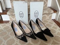 Two pairs of black suede pointed-toe heels displayed side by side with original white Emma Hope's Shoes boxes in background