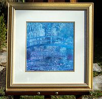 Framed print shown outdoors on easel. Clear view of gold-toned wooden frame, cream double matting, and the blue impressionist water lily bridge artwork.