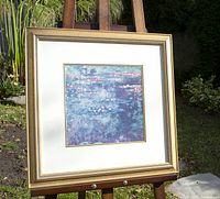 Full view of the framed print on an easel outdoors, showing the gold-trimmed solid wood frame, double white matting, and the square print of water lilies in greenish-blue and purple hues.