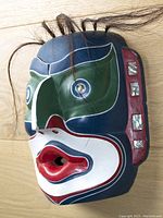 Three-quarter angle of mask showing carved facial features, paint colors and mother of pearl eye inlays