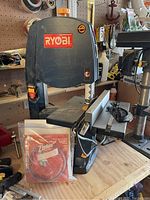Dark gray/black Ryobi bandsaw on workbench with extra sealed bandsaw blade in front.