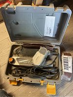 Open light grey plastic carrying case containing electric belt sander with power cord, sanding belts, and manual.