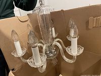 Crystal chandelier showing five lit candle-style bulbs, decorative etched glass arms, and central column. Stored in cardboard box.