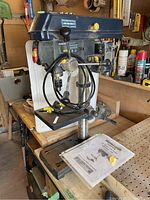 Mastercraft 10-inch bench drill press positioned on a workbench, showing the head unit with laser line, adjustment handles, and included user manual on base.