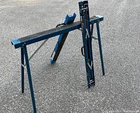 Blue folding Mastercraft sawhorse set with collapsible metal legs and handles for carrying shown unfolded on a paved surface.