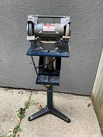 Full view of the grinder sharpener mounted on a heavy-duty metal stand showing the base, motor housing, wheels, and power cord.