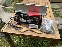 Side view of benchtop jointer mounted on black cast iron base with aluminum tables and red cutterhead guard, wooden push stick and ziplock bag with hardware on wooden deck