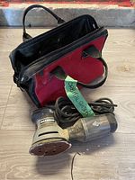 Maximum corded random orbital sander shown with cord and attached sanding pad, placed beside the empty Husky toolbag on a wooden floor.