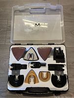 Clear plastic accessory case opened showing assorted oscillating tool blades and sanding pads organized in white insert.