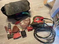 Image of Skil detail sander with power cord, multiple sandpaper attachments in plastic sleeves, and black fabric toolbag laid out on a wood floor.