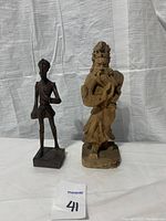 Two Asian wood carved figures placed on white fabric background, one dark slender figure on a square base, one larger light wood figure.