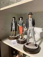Photo showing three ceramic figurine statues of a man and two women on shelves in natural light.