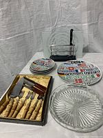 Wide view of all items in the lot on a table including wooden cheese board with bamboo forks and knives, small and large serving plates, clear glass platter, and glass cutting board with slicing wire.