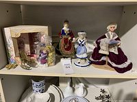 Shelf with four collectible dolls, a brass mini rocking chair, a small doll in packaging, and a floral-patterned box