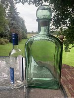 Front view of large green glass decorative bottle with glass stopper and cork next to standard wine bottle to indicate size.