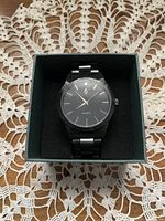 Men's black metal watch in original box on crocheted white doily. Watch face is black with silver hands and markers.