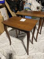 Two wooden nesting tables with medium brown varnish, positioned side by side on a drop cloth in a storage space