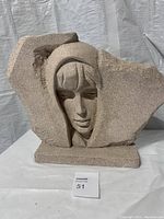 Front view of the heavy concrete or ceramic sculpture showing the stylized hooded female face emerging from an irregular shaped background slab on a rectangular base.