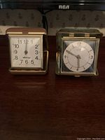 Two travel clocks placed side by side on a wooden surface, showing the front dials. Brown clock on left and green on right.