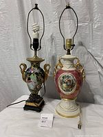Pair of Victorian style table lamps, showing full front view including bases and harp. One lamp black floral on wood base, the other pink and white with gold handles and detailed painting.