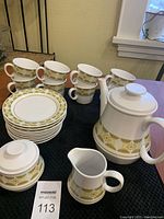 Full view of tea pot, cream and sugar bowl, 8 coffee mugs stacked, and 9 small saucers arranged separately on black cloth.