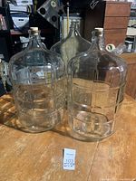 Photo showing three large clear glass demijohns with cork stoppers and cooler style horizontal ridges, placed on wooden table.