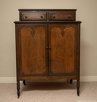 Front view of antique wooden dresser cabinet showing two upper drawers with brass knobs and lower double doors with decorative wood panels and round wooden knobs. The dresser is supported on turned legs with caster wheels.