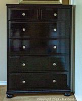 Front view of tall dark wood dresser showing five drawers with round metal knobs, standing on rounded feet.