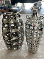 Photo of two decorative silver color vases, one with a grid of nine square holes and one with numerous circular holes and two small handles, both with shiny metal finishes.