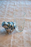 Photo showing a cocktail mixer consisting of a clear glass with etched palm trees and a stainless steel shaker top lying next to it.