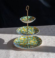 Three tier serving plates with bright floral pattern on each level, mounted on a brass stand with a loop handle on top, photographed against a lace tablecloth and dark background.