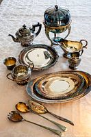 Silver serving items including teapot, hinged dish, gravy boat, creamer, sugar bowl, two oval trays, two serving spoons, and goblet arranged on a tablecloth showing tarnish