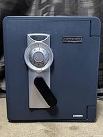 Front view of a dark gray First Alert office safe with a large silver combination dial above a black handle, showing in excellent condition and placed on a carpeted floor against a dark patterned curtain.